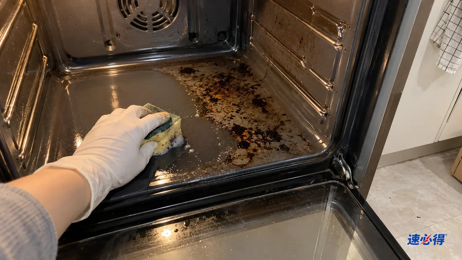 一只戴白色丁腈手套的手用湿海绵擦烤箱底部有光泽的珐琅面，擦过的一块露出干净底色，旁边还有烤焦油渍，侧壁灰色涂层板没有碰。