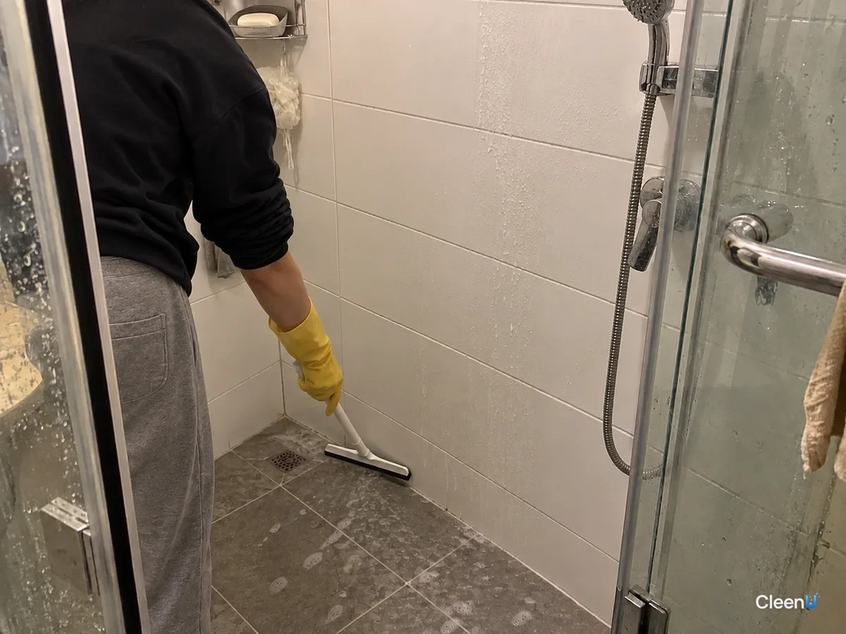 Same shower: the yellow-gloved hand pushes standing water across the tray toward the drain with the squeegee; the wall tile is already done and a small stream gathers at the drain.
