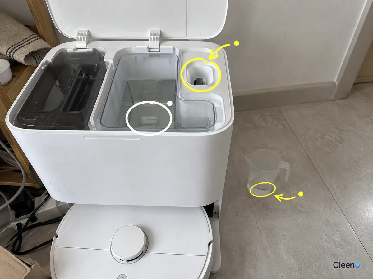 An open robot dock with the auto-dosing solution inlet, the clean tank's outlet and a measuring cup marked for refill design.