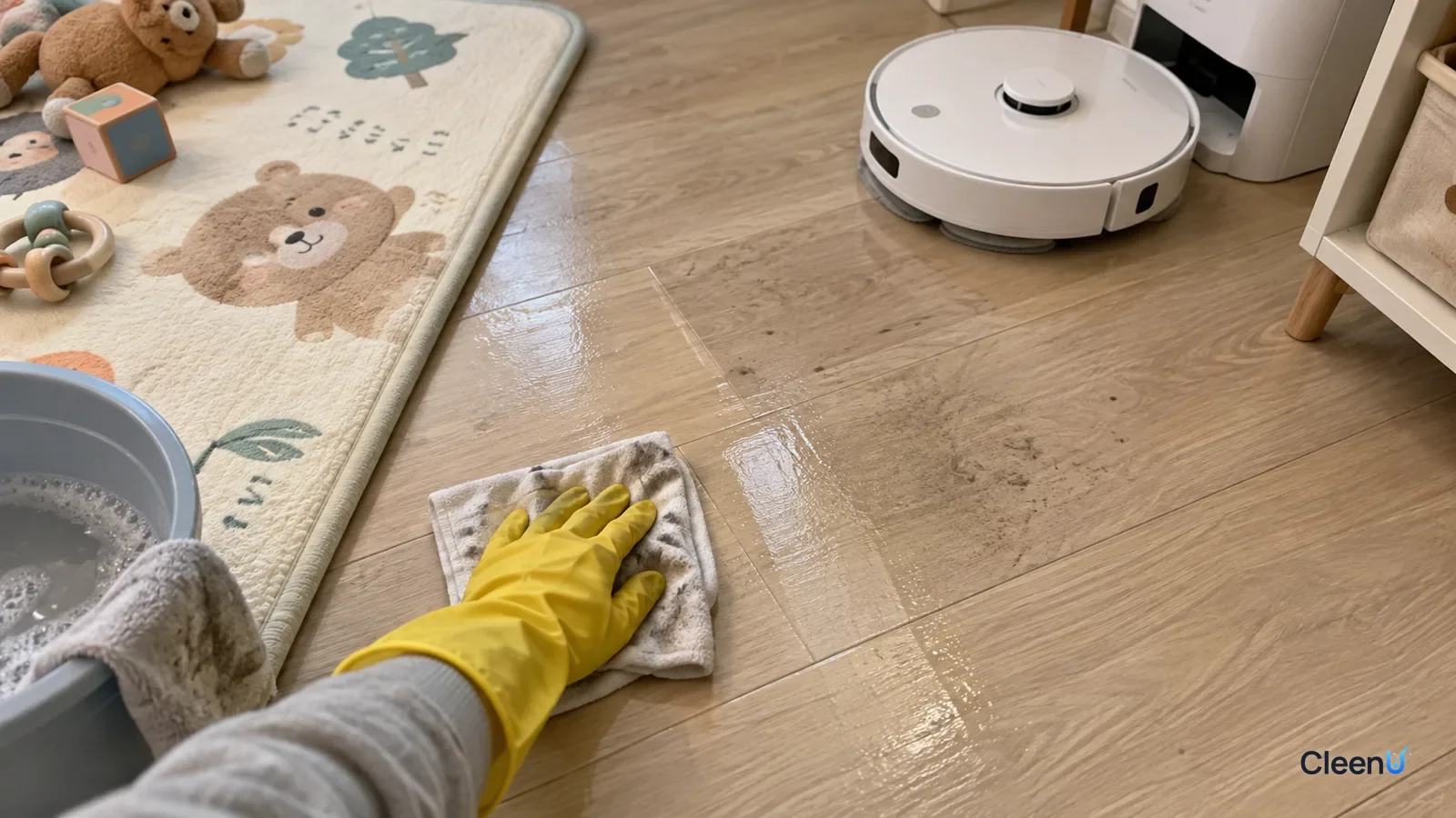 A yellow-gloved hand wipes a section of wood floor with a damp cloth beside a baby play mat while the robot mop works nearby.