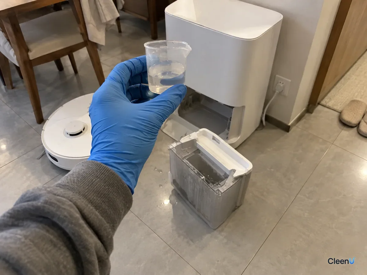 A blue-gloved hand holds a small clear measuring cup up to read its fill marks beside an open white robot mop dock.