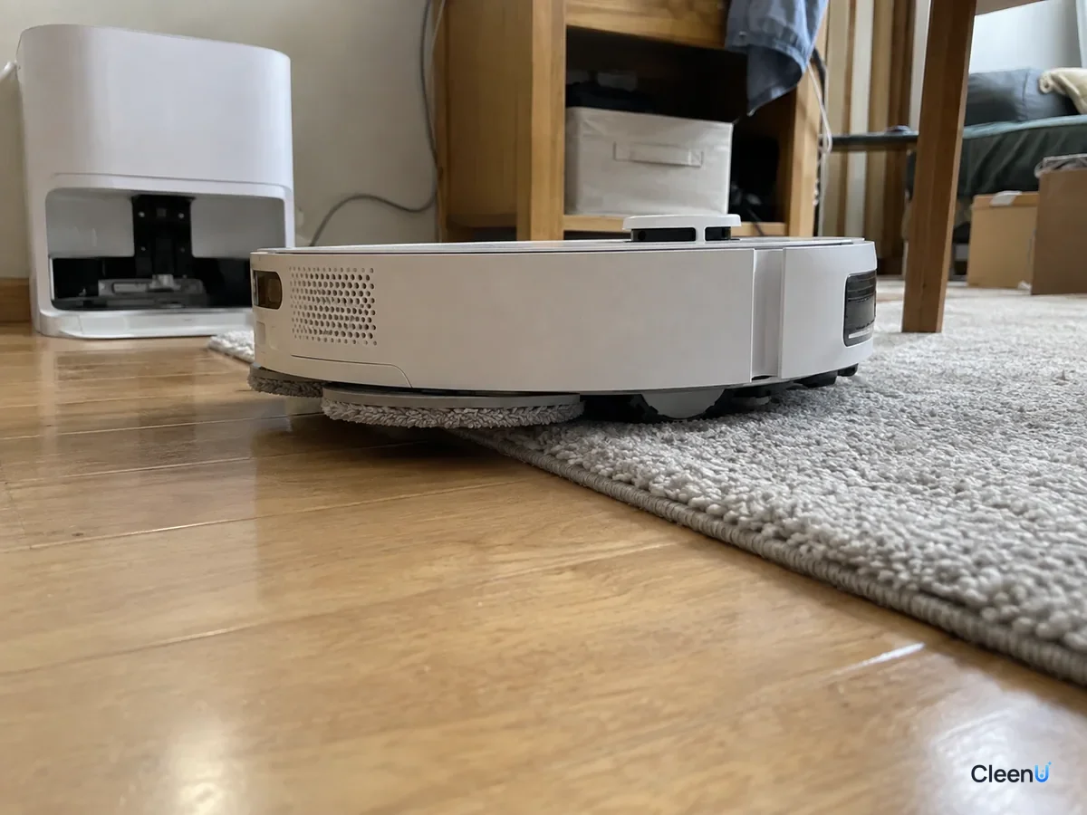 AI illustration: Low close-up of a robot mop with its pads lifted off the floor, rolling from hardwood onto a light gray low-pile rug with a dry edge