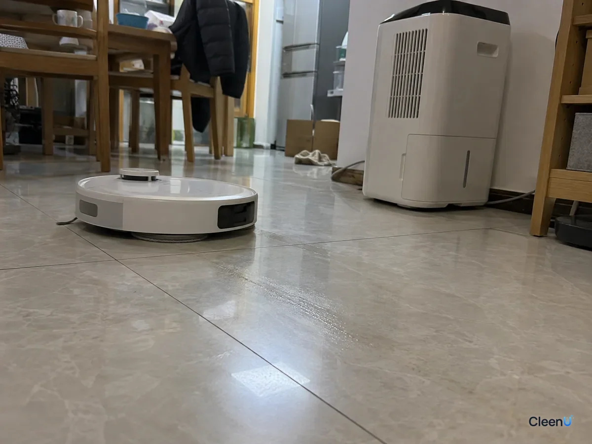 AI illustration: Low close-up of a robot mop on its lowest water setting leaving only a thin damp trail on tile that fades quickly, with a dehumidifier running nearby
