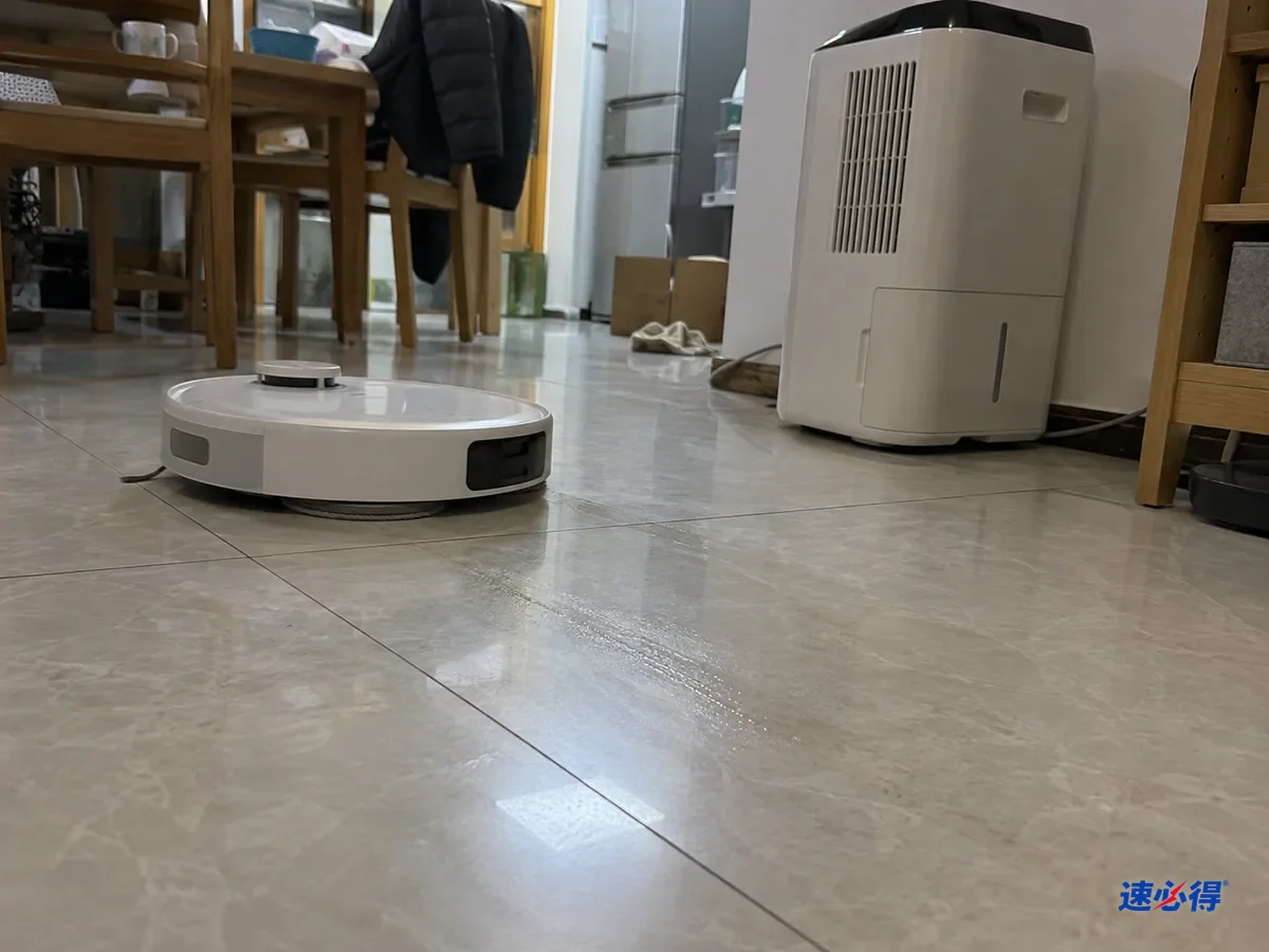 AI 示意图：低机位近景，扫地机器人在出水量最低的状态下拖过瓷砖，地面只留下一层很快消失的薄湿痕，旁边的除湿机正在运行