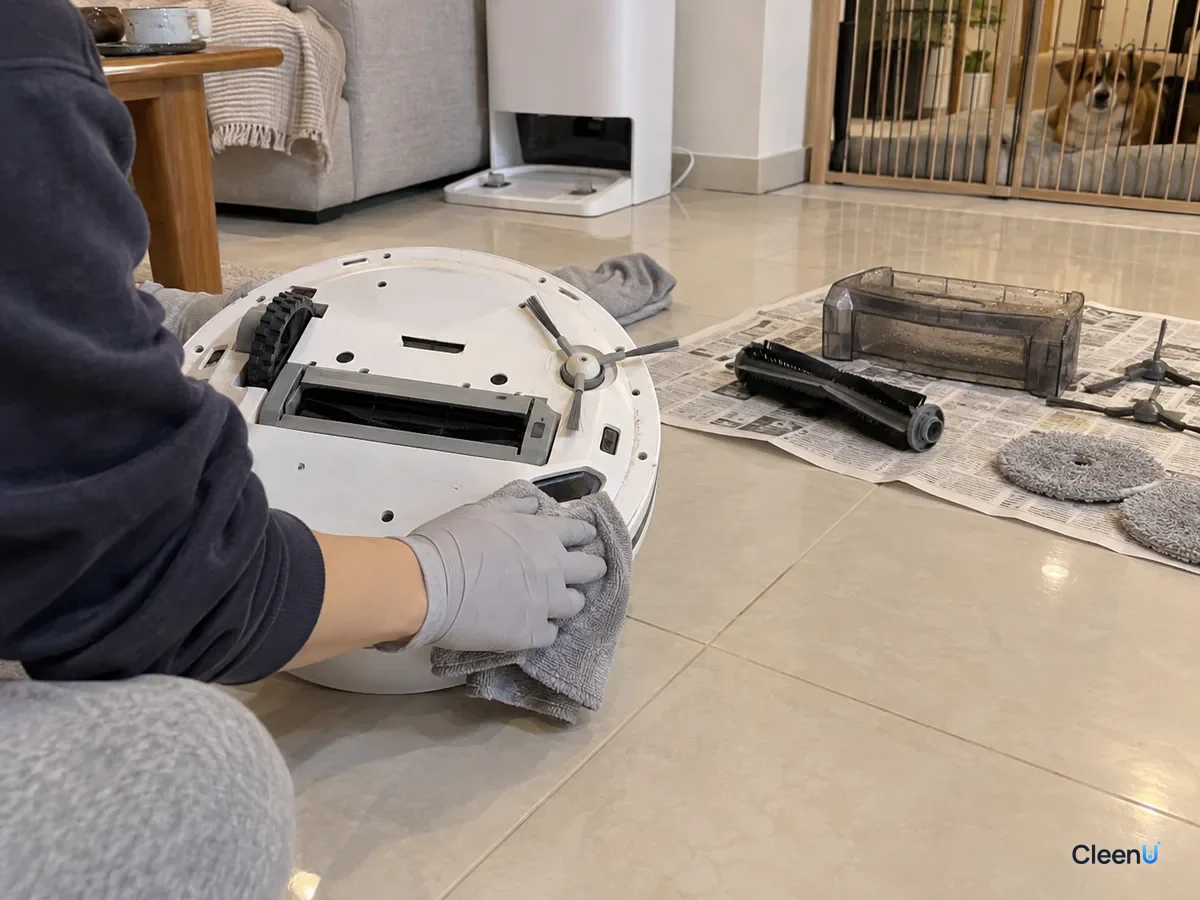 A gloved hand wipes the underside of the robot vacuum with a cloth after pet mess, its removed parts drying on newspaper.