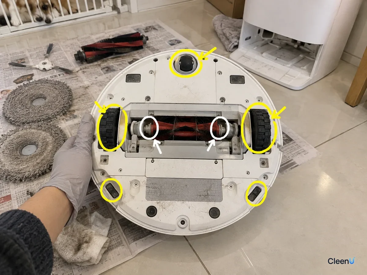 An overturned robot vacuum with the front wheel, both drive wheels, roller brush ends and charging contacts marked for cleaning.