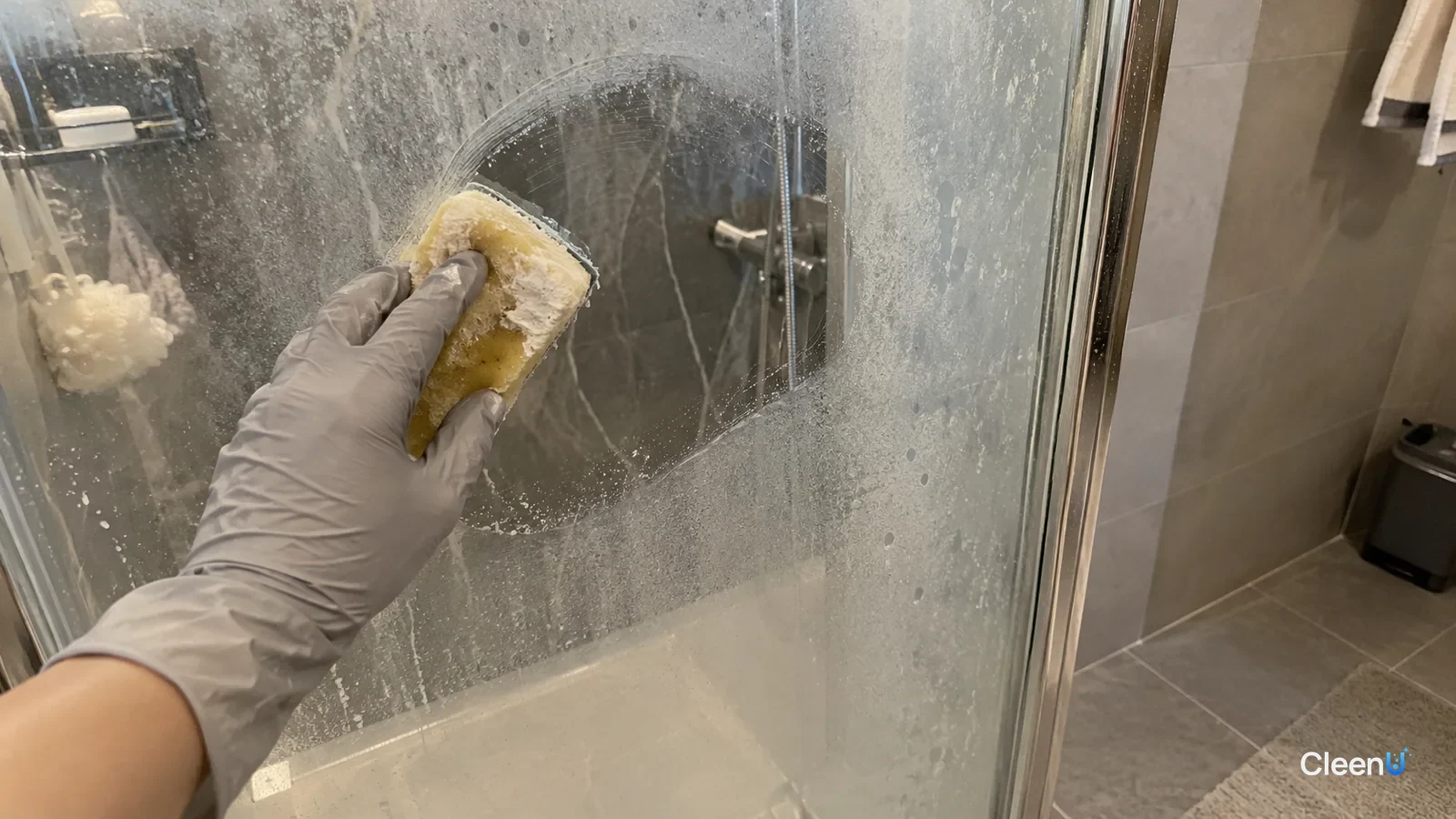 First-person: a light gray nitrile-gloved hand circles a cloudy shower door with a soft sponge, one clear round patch, white residue on the sponge.