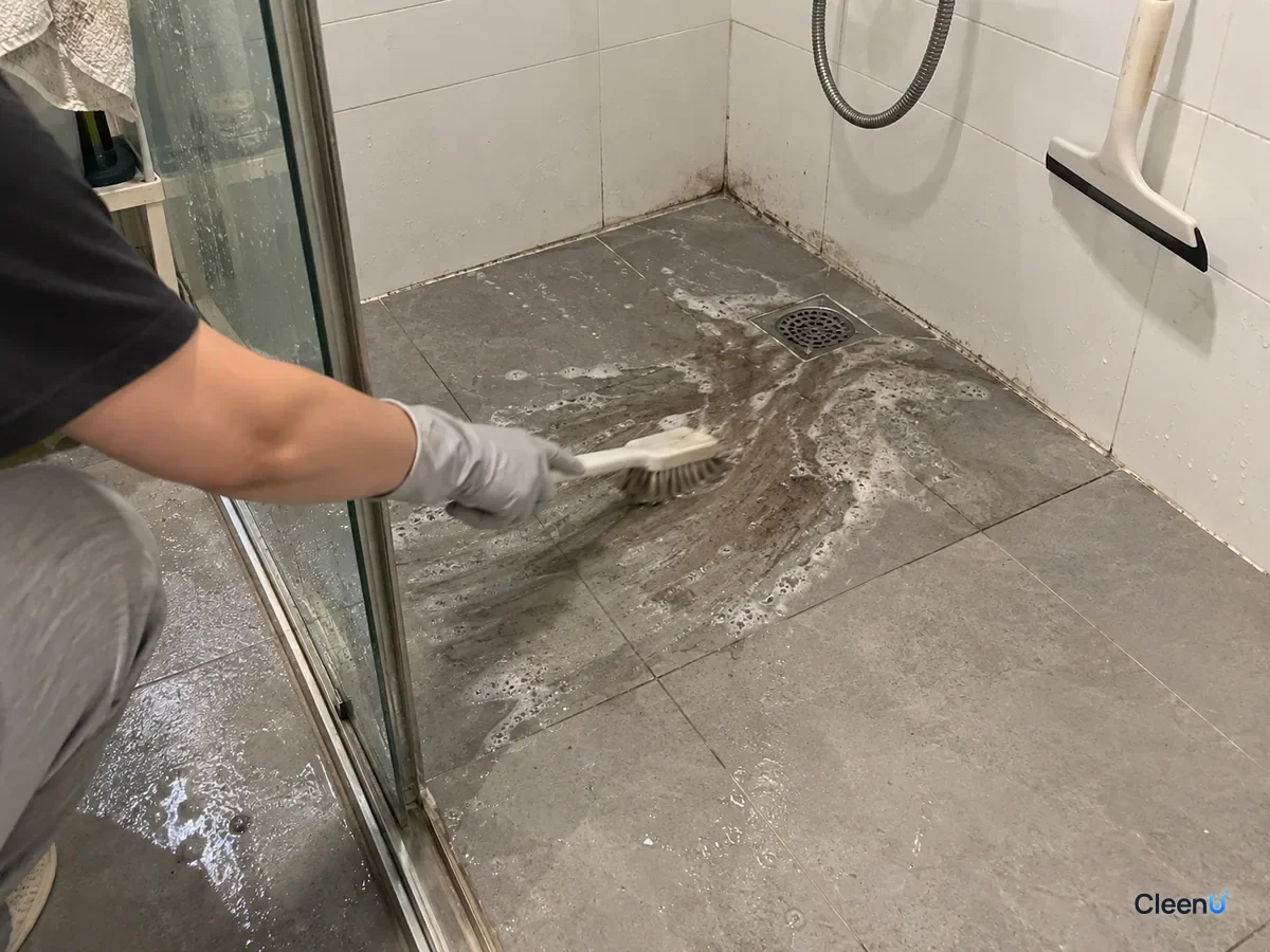 Light grey nitrile-gloved hand scrubbing the anti-slip shower floor tiles with a floor brush, grey-black murky water flowing toward the drain, the corner not yet done