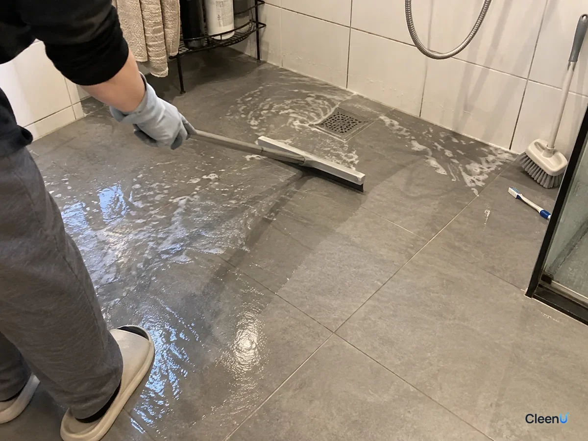 Same bathroom floor later: grey-gloved hand pushing rinse water into the drain with a floor squeegee, the scraped tiles already turning dry