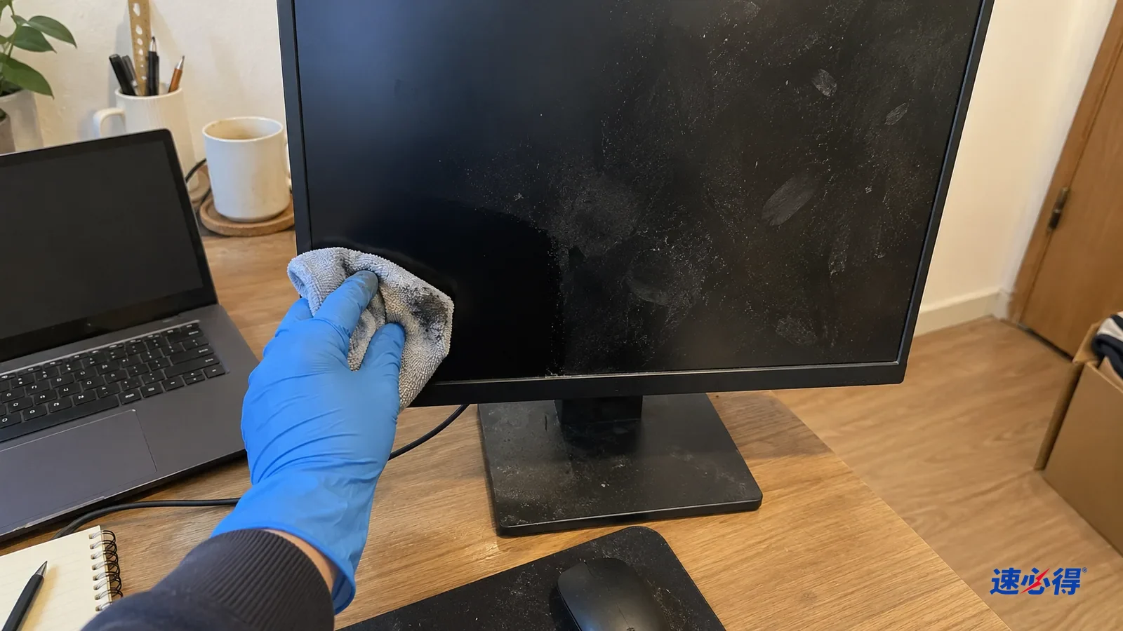 一只戴蓝色丁腈手套的手用微湿超细纤维布轻擦关着的显示器左下角，布面沾灰，擦过的一块黑净，旁边还有干水点和指纹。