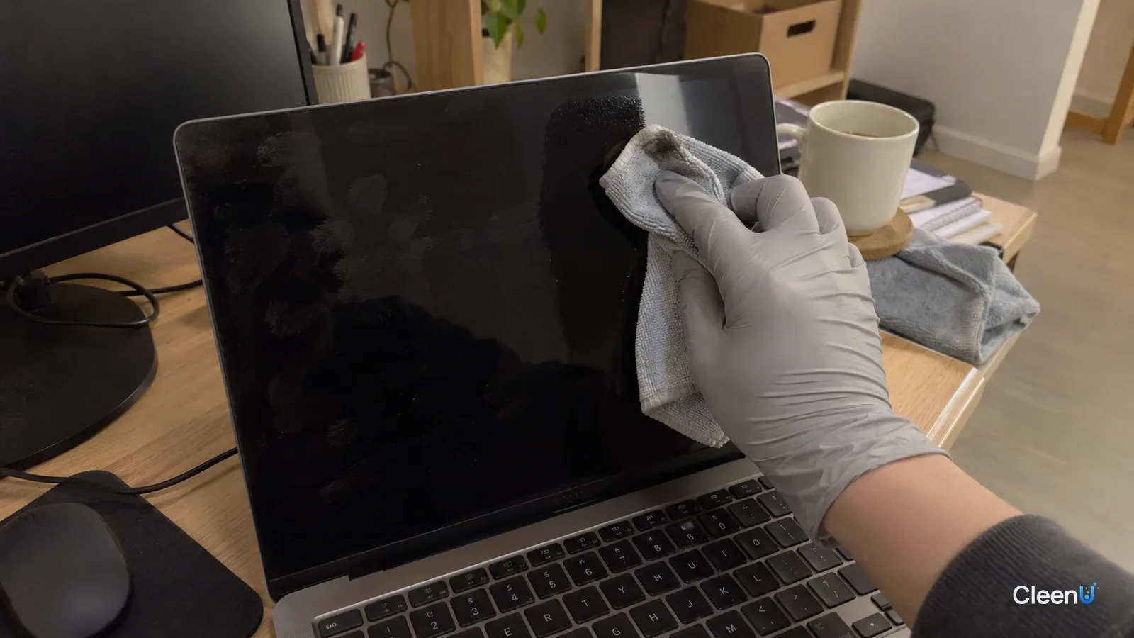 First-person close-up: a hand in light gray nitrile gloves wipes the edge of a switched-off laptop screen with a damp microfiber cloth corner; faint gray on the cloth, fingerprints on one side.