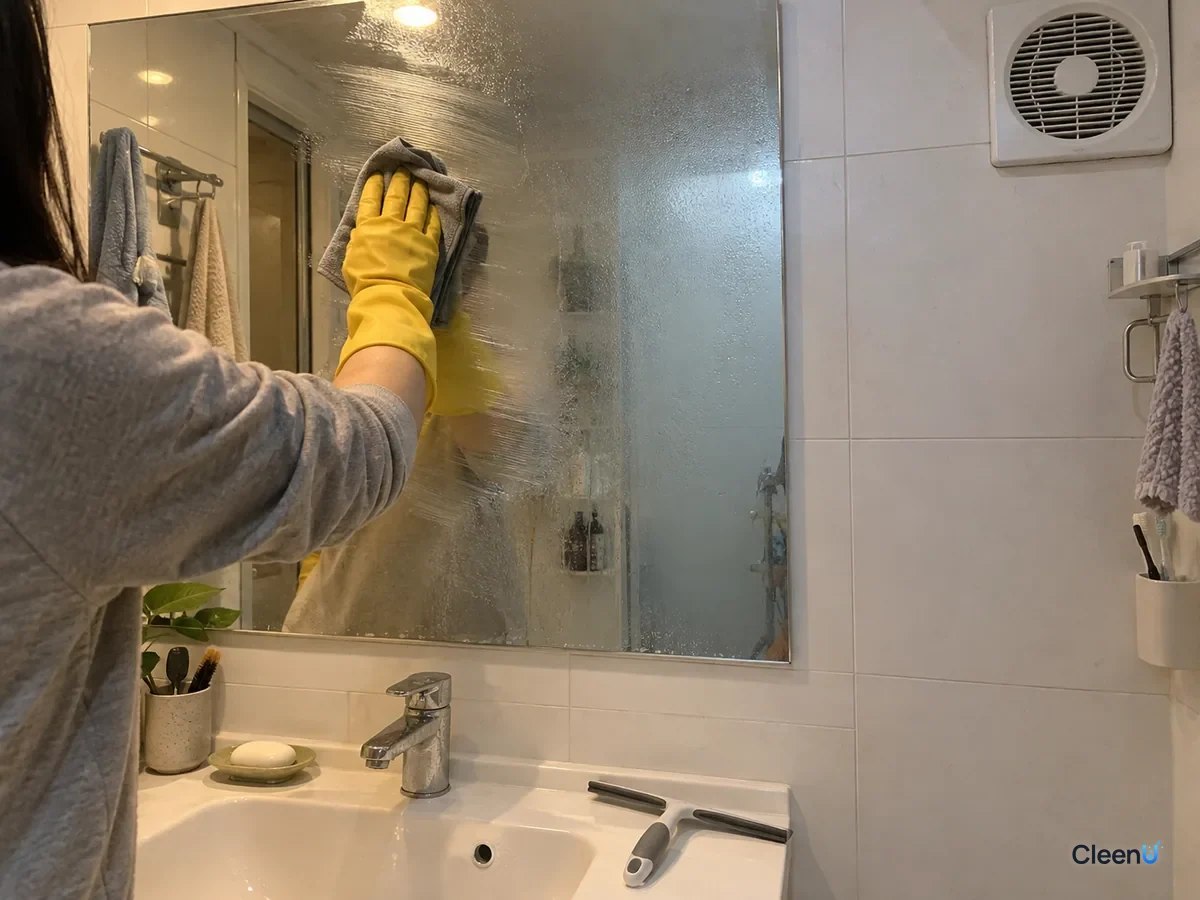 From a step back: a yellow rubber-gloved hand buffs thin anti-fog onto half the mirror with a dry microfiber cloth, faint wet streaks remain, the other half untreated, fan grille in the corner.