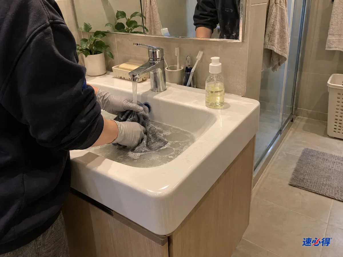 浴室洗手盆边一步外看，戴浅灰色丁腈手套的手在温水里搓洗超细纤维布，盆里水微微发灰，旁边一小瓶中性洗涤剂。