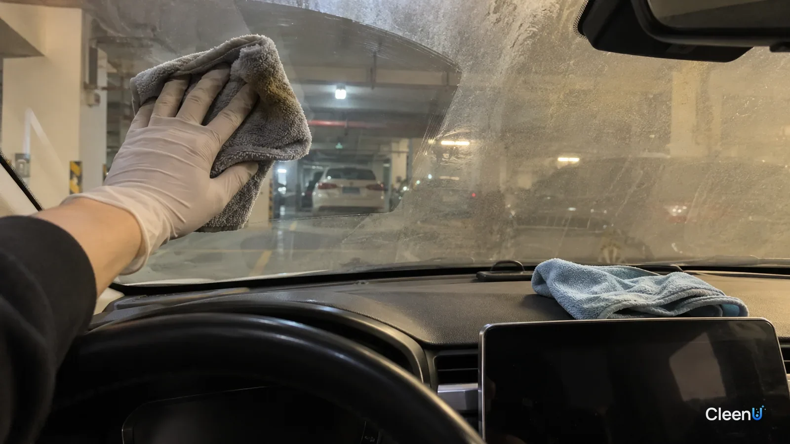 In a parked car, white nitrile-gloved hand wiping the inner windshield with microfiber, greyish-yellow film on the cloth, one patch clear, the lower half still hazy