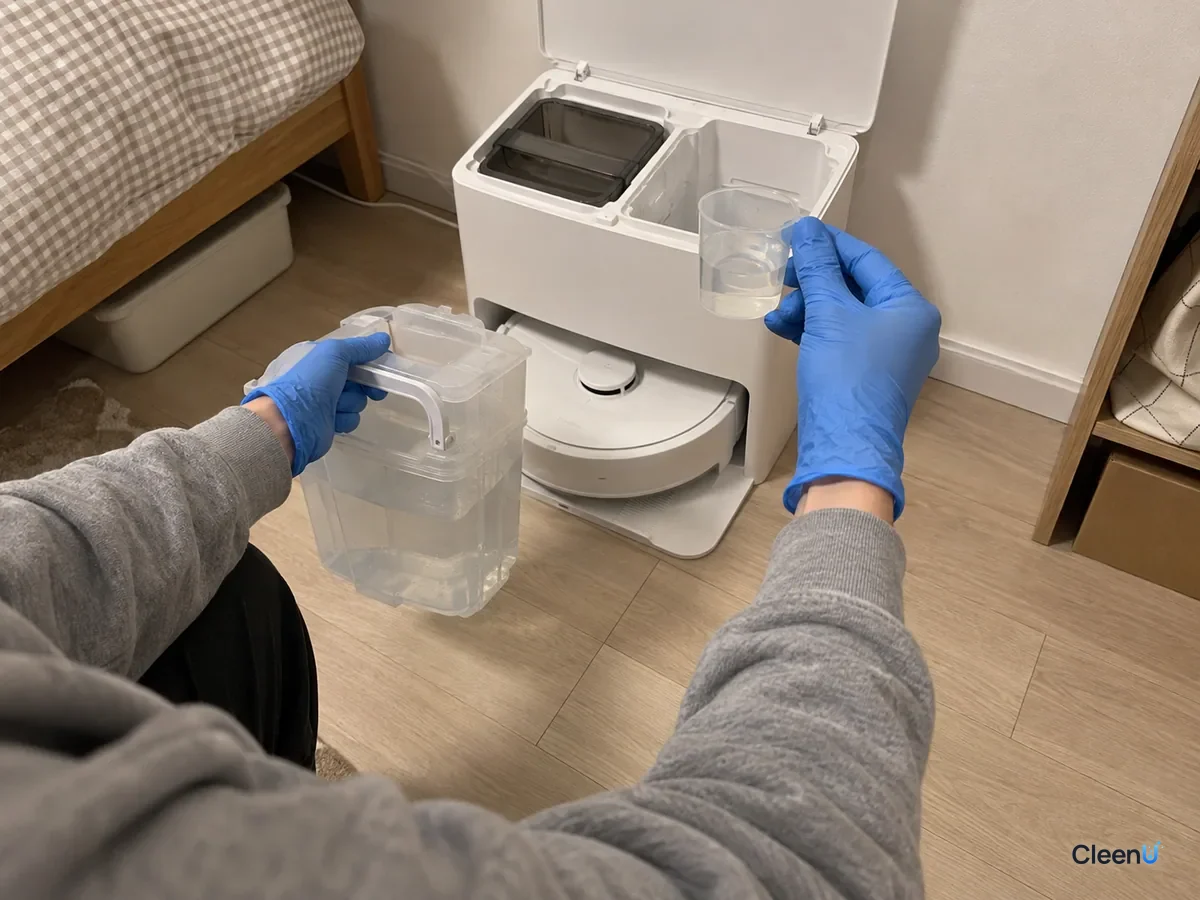 A blue-gloved hand holds a filled clean tank above the floor, with a small measuring cup in the other hand, by the open base.